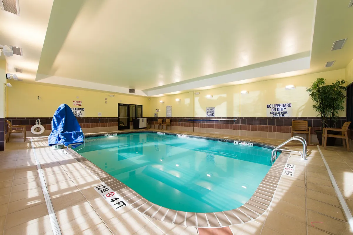 A bright and inviting indoor pool area with a blue pool cover, wooden chairs for lounging, and lush indoor plants. The setting is ideal for both peaceful solo swims and lively group activities, reflecting a tranquil, refreshing mood.