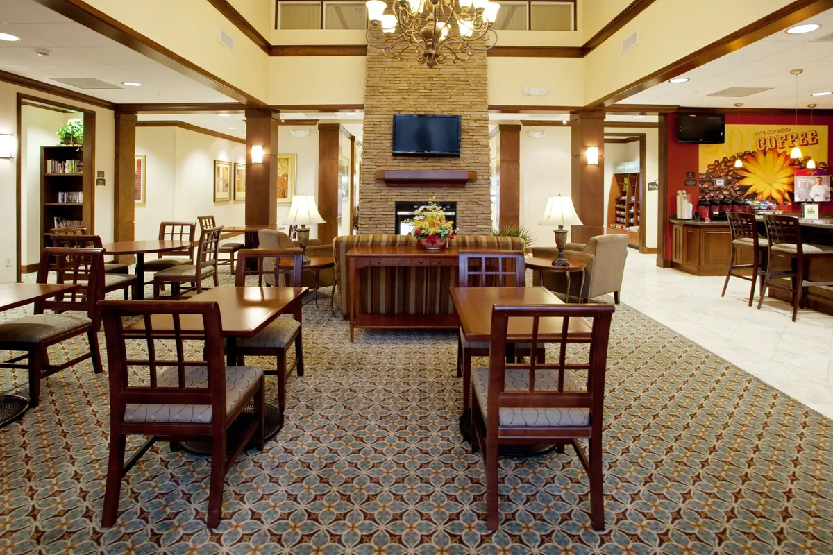 A bright, social space showcasing a central fireplace, wooden tables and chairs, a chandelier, and a well-stocked coffee station. The scene suggests guests enjoying coffee, reading, and gathering by the fire, creating a sense of warmth and community.