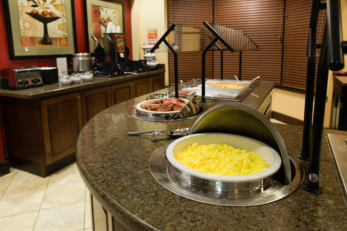 A bright buffet counter displays yellow scrambled eggs, a meat tray, and a well-stocked coffee station. The scene captures a casual dining setup, with inviting lighting and a clean, organized food presentation.