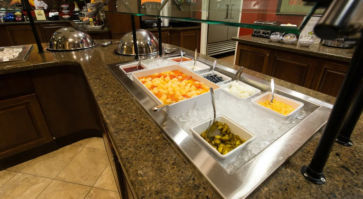 A vibrant display of fresh fruit salad, toppings, condiments, and an ice display suggests a self-serve dining area inviting guests to create personalized plates. The casual, friendly setting encourages guests to socialize as they make food choices together.