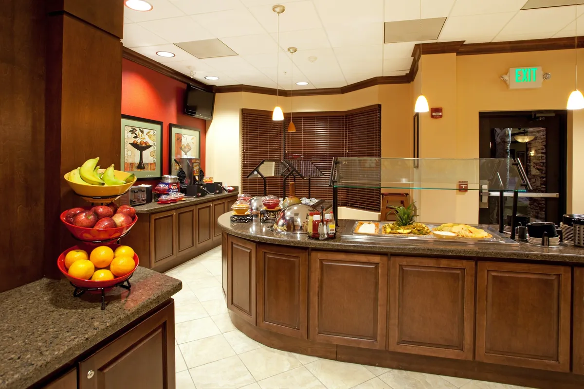 A welcoming studio living space with a cozy breakfast bar, a colorful fruit bowl, and guests enjoying breakfast or socializing. The scene is filled with natural light, suggesting a cheerful and inviting ambiance.