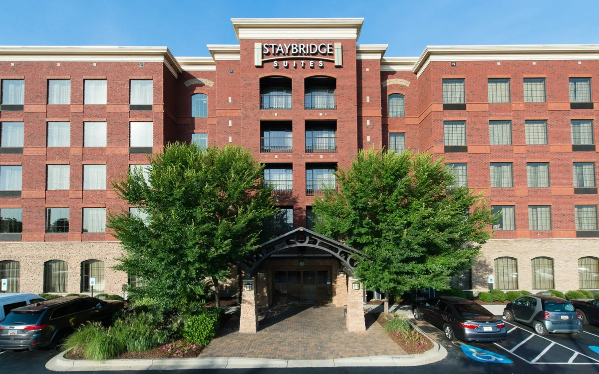 A welcoming exterior view of the Staybridge Suites entrance, showcasing the brick facade, lush trees, and a spacious, convenient parking lot for guests.