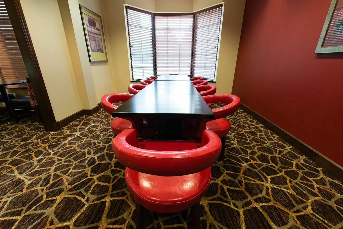 This image features a long black table surrounded by striking red circular chairs, set against the backdrop of large windows that allow plenty of natural light into the space. The area is designed for social gatherings and shared meals, with a dynamic and contemporary feel that encourages group interaction and conversation.